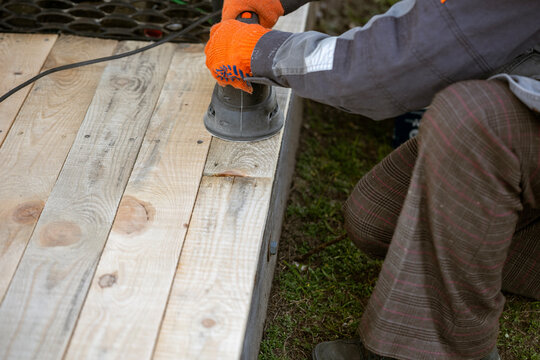 A worker in an orange glove uses an electric orbital sander to smooth new, light-colored wooden planks. This DIY woodworking project involves preparing a deck or floor surface outdoors.