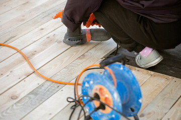 A worker in an orange glove uses an electric orbital sander to smooth a new wooden deck. An orange extension cord and a blue cable reel are on the planks.