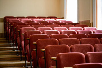 A side view of an empty university lecture hall. Rows of maroon seats with small wooden desks are arranged on stepped risers, with bright windows in the background.