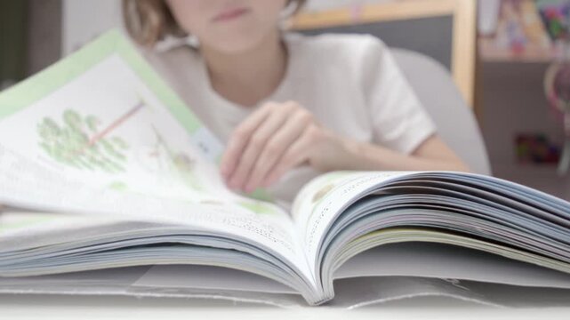 Child in white t-shirt flipping pages of book at desk, exploring new information with focus and curiosity, close-up view of reading process