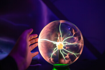 A hand touching a plasma ball in the dark. The bright purple and magenta electrical filaments follow the fingertips, demonstrating physics, static electricity, and energy in an interactive lamp.