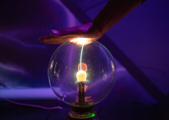 A single, bright filament of electricity jumps from the central electrode of a plasma ball to a hand hovering above, demonstrating static interaction in a dark room.