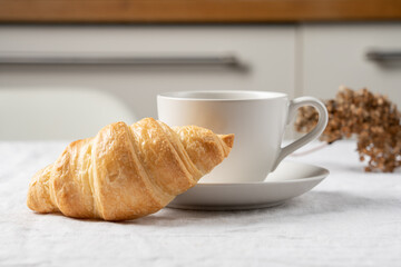 Fresh croissant and cup of coffee on table with tablecloth, cozy breakfast and morning coffee concept