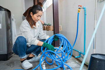 Asian woman in casual outfit wearing gloves is organizing blue garden hose at home while practicing sustainable gardening and eco lifestyle for better living and home care