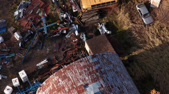 Drone video showing a cluttered scrapyard with rusted metal parts, industrial debris, an overgrown vintage bus, and an abandoned car among tall grass and scattered materials.