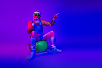 Retro bearded fitness man in neon purple and blue lighting points forward while sitting on a green exercise ball in a retro sporty studio