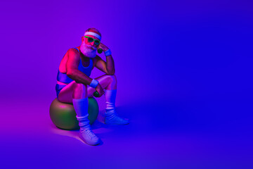 Fototapeta premium Neon retro fitness man sits on an exercise ball under purple and blue lights with bold style energy and joyful attitude