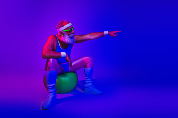 Energetic elderly man in retro fitness outfit sits on a green exercise ball and points forward against a neon purple and blue backdrop
