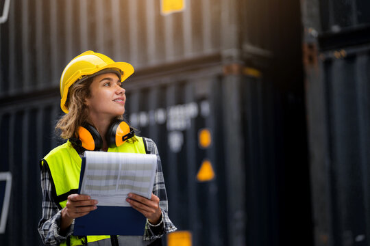 A woman wearing a yellow vest and a hard hat is holding a clipboard