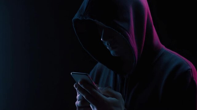 Digital Shadow: Enigmatic figure shrouded in a hooded sweatshirt, engrossed in the glow of a smartphone. A shadowy scene evoking intrigue and the clandestine aspects of the digital realm.