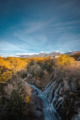 Fototapeta premium Vertical Japanese autumn view in the mountains with a blue water, yellow trees, and a partly cloudy sky at Shirahige Waterfall, Hokkaido, Japan