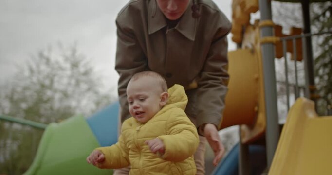 A mother helps her toddler down a slide at the playground. The child is wearing a yellow jacket and is enjoying the activity.