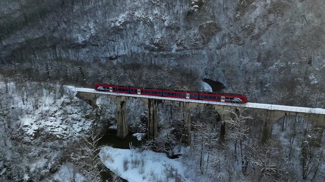 Aerial view of a red and blue train cutting through a snow-covered landscape, contrasting against the white surroundings, Valjevo, Kolubarski okrug, Serbia.
