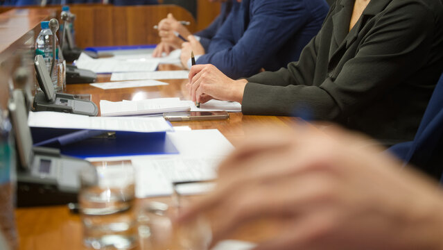 Close-up of anonymous hands in suits holding pens over documents at a board meeting; smartphone, folder and mics on the wooden table, selective focus. Photo