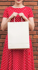 Stylish woman in a retro red polka dot dress holding a blank white paper shopping bag for a mockup against a brick wall background