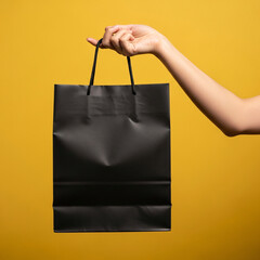 A person's hand holding a plain black paper shopping bag against a solid yellow background. Concept for retail, sales, or gift-giving