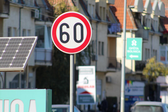 Close-up of a 60 km/h speed limit sign next to a solar panel in a city street with residential buildings in the background.