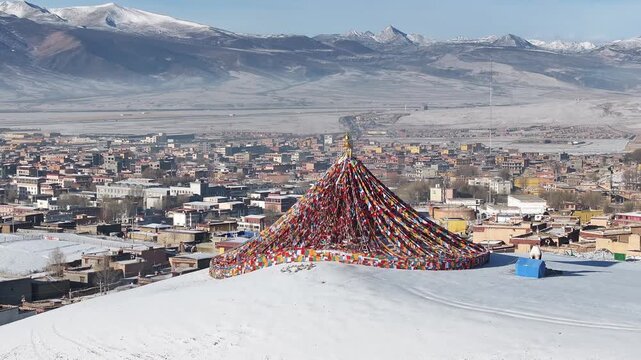Aerial view of prayer flags cascading down a snowy hill with the city of Litang and snow capped mountains in the background, Litang, China.