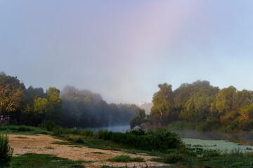 Fototapeta premium Misty river unveils a tranquil, enigmatic morning. Gentle fog hugs the shore, creating a serene, peaceful landscape with soft light. Nature's quiet awakening