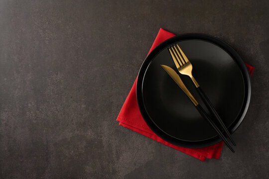 Minimalist table setting with a matte black plate and gold cutlery resting on a red napkin against a dark textured surface, evoking festive Christmas and New Year ambiance for holiday dining