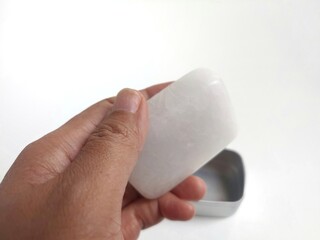 A hand holding a natural alum mineral crystals in square shape or tawas as underarm deodorant with reusable metal packaging isolated on white background
