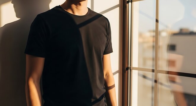 A model stands by a window wearing a blank black t-shirt in golden hour light.