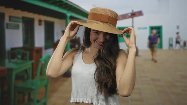 Woman adjusts straw hat while smiling with long dark hair in white tank top against colonial building; joy confidence.