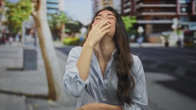 Young woman with long hair covering mouth with hand while smiling on busy city street during daytime; laughter.