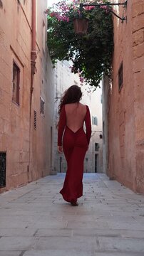 A woman in a backless red gown walks away in a narrow limestone alley in Valletta, Malta. Soft daylight, leading lines, pink bougainvillea, warm stone tones.