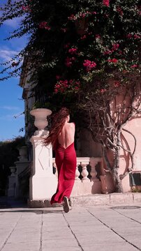 A woman in a backless red gown walks and poses by a sunlit limestone balustrade in Valletta, with pink bougainvillea, strong contrast, and a romantic editorial mood.