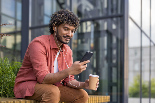 Smiling young Indian man sitting on a bench outdoors wearing headphones, using mobile phone and holding a cup of coffee