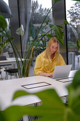 Gen Z freelancer with laptop working in a green coworking space among indoor plants, biophilic...