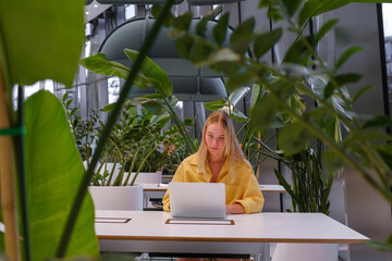 Gen Z freelancer with laptop working in a green coworking space among indoor plants, biophilic design; remote work, focus, productivity, flexible schedule, sustainable work lifestyle.