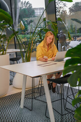 Gen Z freelancer with laptop working in a green coworking space among indoor plants, biophilic design; remote work, focus, productivity, flexible schedule, sustainable work lifestyle.