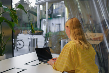 Gen Z freelancer with laptop working in a green coworking space among indoor plants, biophilic design; remote work, focus, productivity, flexible schedule, sustainable work lifestyle.