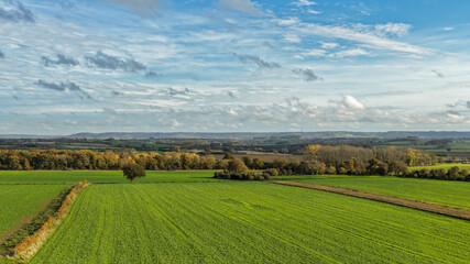 Aerial view of green farmland fields with tree-lined hedgerows, distant hills and a bright blue...