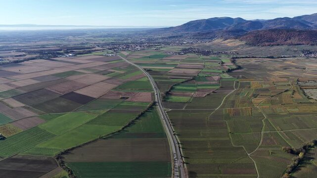 Aerial view of Route D83 cutting through a patterned landscape, a tapestry of fields and vineyards, under a clear sky, Rouffach, Grand Est, France.
