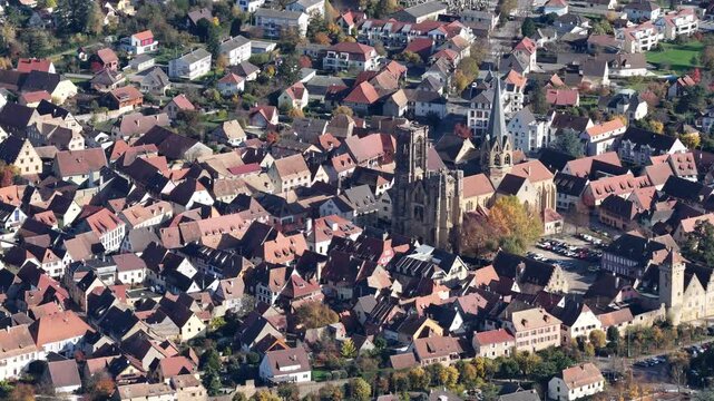 Aerial view of buildings with red rooftops, a church with a tall spire, and greenery casting shadows across the town, Rouffach, Grand Est, France.