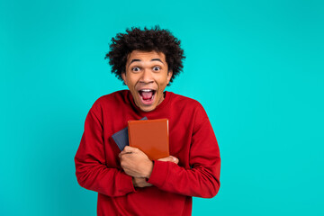Young man in red sweater holds books and shows excited joyful expression against teal background