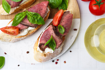 Delicious bruschettas with roast beef served on white tiled table, flat lay