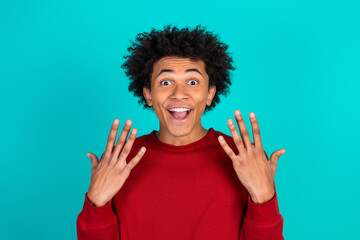 Cheerful young man in red sweater shows excited expression with open hands against a vibrant teal background