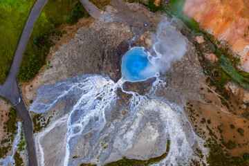 Aerial top down view shows a turquoise hot spring venting steam in Iceland, mineral streaks and white silica runoff fan outward, bright daylight reveals crisp textures.