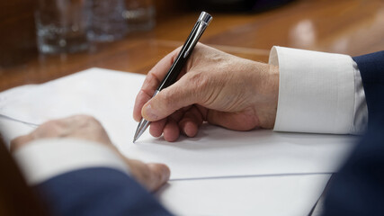 Close-up of an elderly businessman&rsquo;s hand in a suit writing with a metal pen on blank papers at a wooden desk; legal document or contract moment in a boardroom. Phot