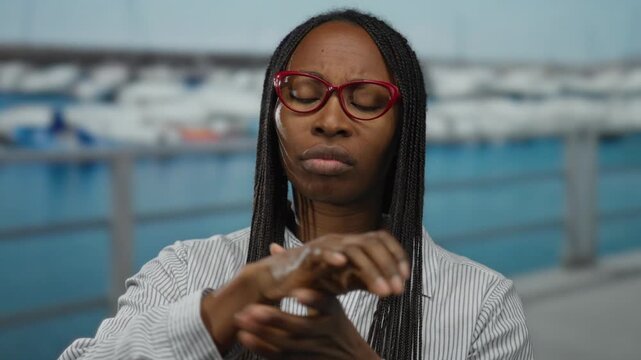 Woman with glasses tending to wrist pain at seaside port with boats in background showcasing outdoor setting and focus on health and relaxation.