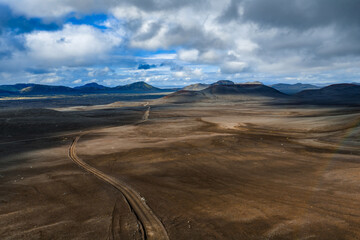 Aerial view shows a barren highland plain of dark volcanic earth in Iceland, a lone dirt track leading to rounded tuya like mountains under diffused daylight.