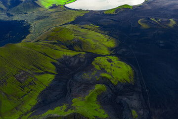 Aerial view of Iceland's Landmannalaugar shows green moss on volcanic ridges, black lava sands, and a pale crater lake in clear daylight with strong contrasts.
