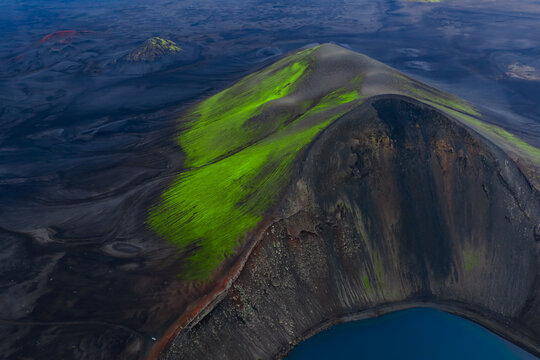 Aerial view of a basaltic crater near Landmannalaugar in Iceland, a turquoise lake at its base, neon green moss on upper flanks, cool low light with long shadows.