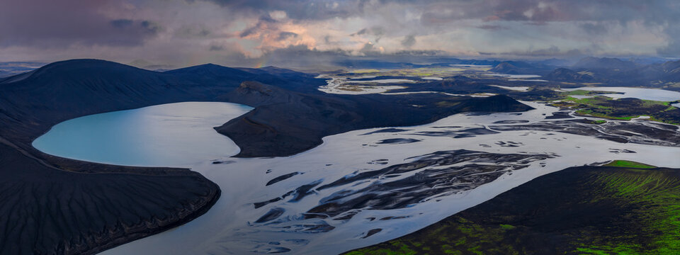 A wide aerial view shows Iceland highlands near Landmannalaugar, a pale blue crater lake by dark ridges, braided glacial rivers, moss patches, and low storm clouds.