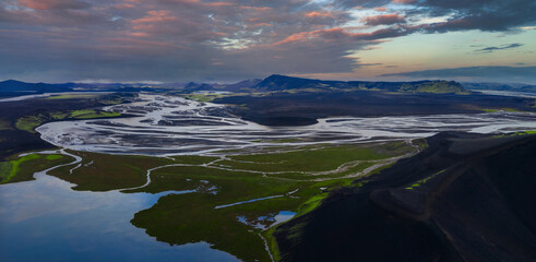 Aerial view shows braided white channels over dark volcanic plains near Landmannalaugar, with green moss, reflective lakes, distant table mountains, and soft evening light.