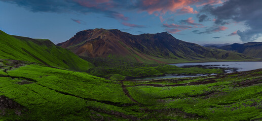 Wide dusk view of Landmannalaugar in Iceland, with green mossy slopes, a ribboned river, a reflective lake, and multicolored rhyolite mountains under soft pink clouds. © Aerial Film Studio